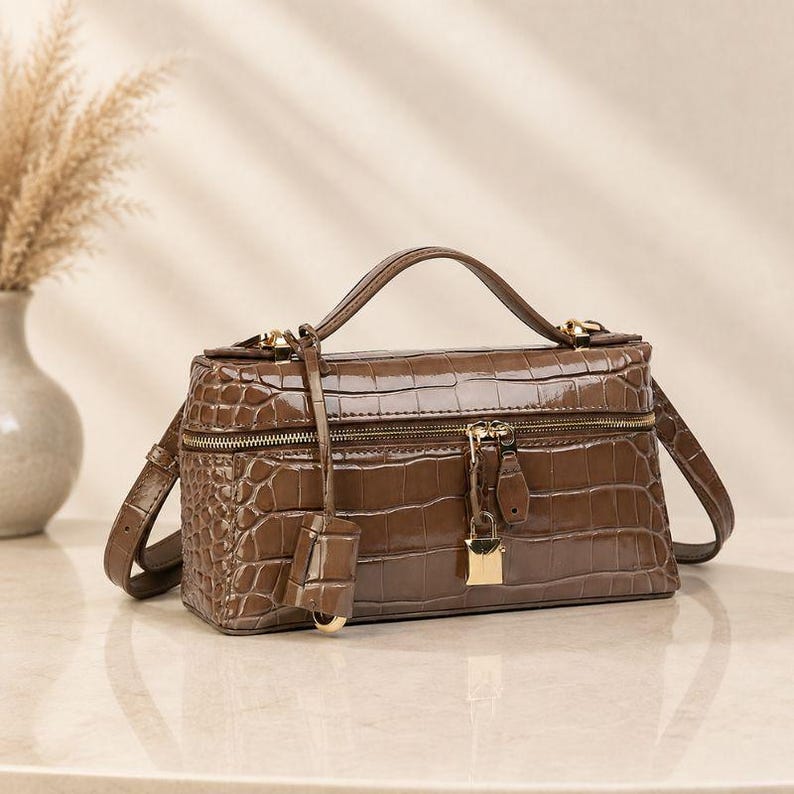May include: A rectangular brown handbag with a glossy crocodile-embossed texture. The bag features a top handle, a detachable shoulder strap, a gold-tone zipper closure, and a small gold-tone padlock hanging from the front zipper pull.