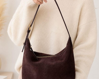 May include: A dark brown suede hobo bag featuring a curved top silhouette, a single adjustable shoulder strap with a gold-toned metal buckle, and a horizontal zipper closure across the top with a matching suede zipper pull tab.