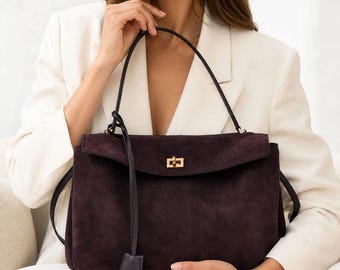May include: A dark purple suede handbag featuring a rectangular silhouette, a fold-over flap with a gold-toned metal turn-lock closure, and a thin leather shoulder strap with a dangling leather tag accent attached to the side hardware.