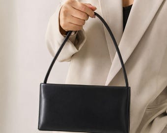 May include: A sleek, rectangular black handbag with a smooth, matte finish and a single thin shoulder strap. The bag features a structured, minimalist design with clean lines and a flat base, providing a compact and functional accessory for daily use.