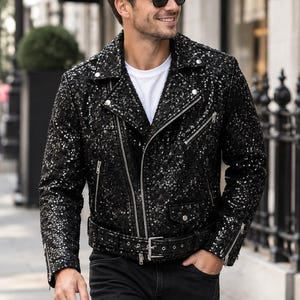 Puede incluir: Una chaqueta de motociclista negra cubierta de lentejuelas brillantes, con solapa de muesca, cremalleras de metal plateado en la parte delantera y los puños, un cinturón con hebilla a juego en la cintura y detalles de botones a presión.