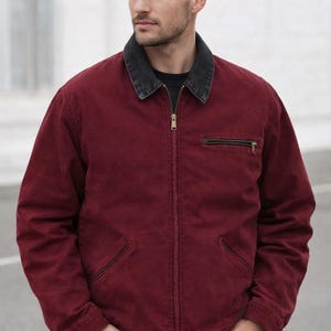 May include: A deep red canvas jacket featuring a dark grey corduroy collar, a full-length front zip, and a horizontal zipped chest pocket. The jacket includes two slanted lower pockets and long sleeves with cuffs, worn over a black shirt and blue jeans.