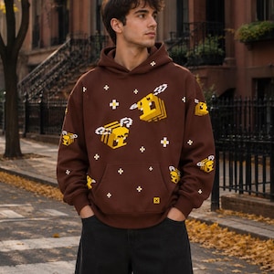 May include: A brown hooded sweatshirt featuring a repeating pattern of yellow pixelated bees and small white cross symbols. The long-sleeved garment includes a front kangaroo pocket and a small yellow square icon near the bottom hem.