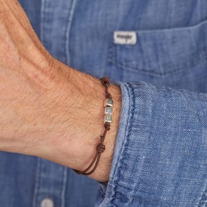Peut inclure: Un bracelet en cordon marron avec trois perles carrées argentées. Le bracelet est noué et porté au poignet d'une personne, contre une chemise en jean bleue. L'étiquette Wrangler est visible sur la chemise.