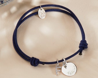 Op de afbeelding: Een donkerblauwe koordarmband met twee zilveren bedels. Een bedel is een klein ovaal met het woord "Elle" erop gegraveerd. De andere bedel is een ronde schijf met het woord "Felicia" erop gegraveerd. De armband heeft ook een zilveren letter "F" bedel met kleine heldere steentjes.