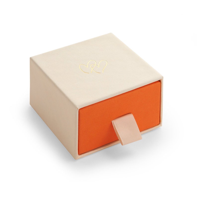 K&ouml;nnte beinhalten: Eine kleine, cremefarbene Schachtel mit einer Schublade. Die Schublade ist orange und hat einen hellrosa Schleifen-Zug. Die Schachtel hat ein goldenes Herz-Design auf der Oberseite.