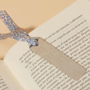 May include: A silver metal bookmark with engraved text, featuring a floral patterned ribbon in blue and white. The bookmark is placed in an open book with French text.