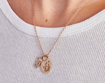 Op de afbeelding: Een goudkleurige ketting met een fijne ketting. De hanger heeft een cirkelvormige bedel met een familiefiguurontwerp en een kleinere bedel met een geometrisch ontwerp. De ketting wordt gedragen over een wit geribbeld shirt.