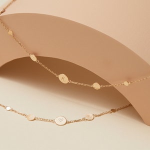 May include: A gold necklace with a delicate chain and small round charms. The charms are engraved with letters and a heart symbol.