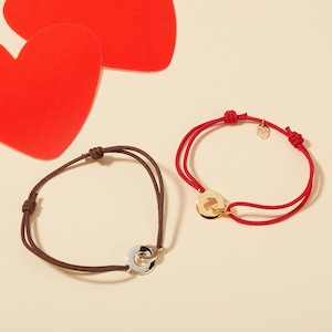 May include: Two adjustable bracelets. One is brown with a silver ring, the other is red with a gold ring. Both have a sliding knot closure. Red paper hearts are in the background.