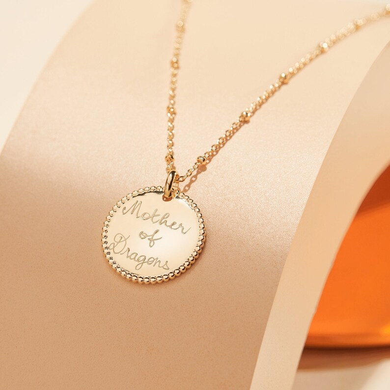 May include: A gold necklace with a round pendant that says "Mother of Dragons" in cursive script.