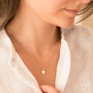 May include: A delicate gold chain necklace with a small star pendant. The necklace is worn by a person with long brown hair and a white blouse.