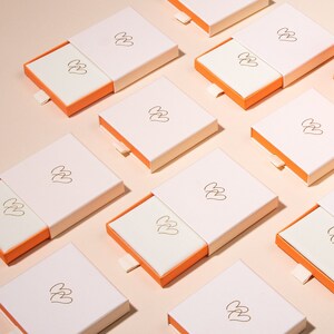 May include: Multiple white boxes with a gold logo of three hearts stacked on top of each other. The boxes have a light orange pull-out drawer.