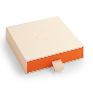 May include: A small, cream-colored box with a drawer. The drawer is orange and has a cream-colored pull tab. The top of the box has a gold design of two intertwined hearts.
