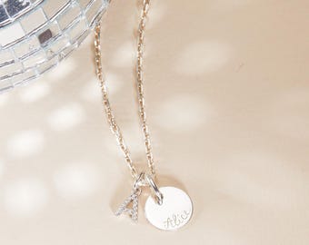 Op de afbeelding: Zilveren ketting met een kleine hanger en een bedel. De hanger is een kleine, ronde schijf met de naam "Alice" in cursief. Een bedel in de vorm van de letter "A" is ook bevestigd. De ketting ligt op een lichtbeige achtergrond.