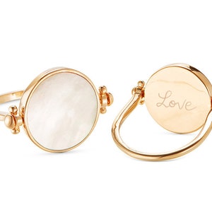 K&ouml;nnte beinhalten: Ein goldener Ring mit einer wei&szlig;en Perlmuttseite und der Gravur "Love" auf der anderen Seite.