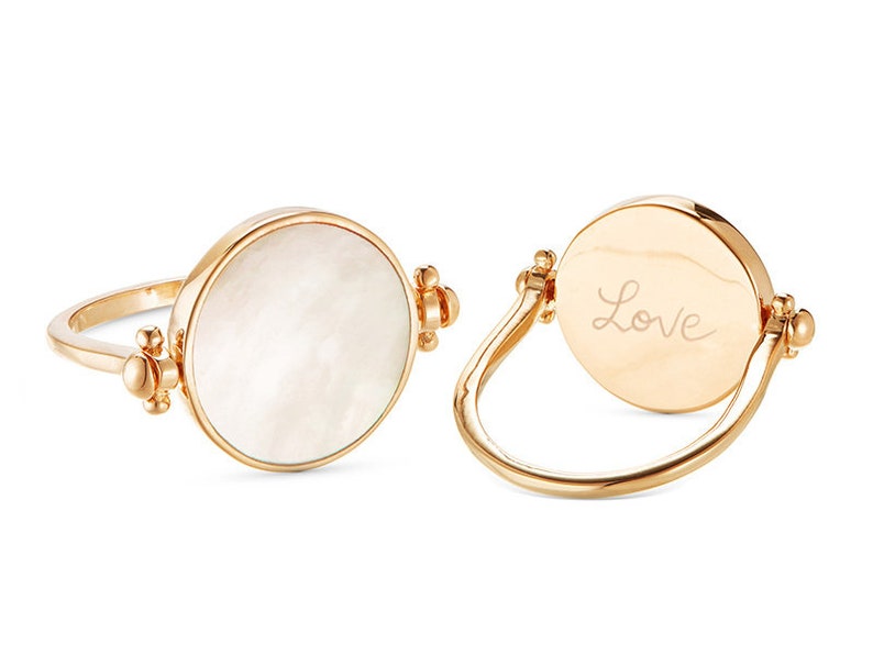 K&ouml;nnte beinhalten: Ein goldener Ring mit einer wei&szlig;en Perlmuttseite und der Gravur "Love" auf der anderen Seite.