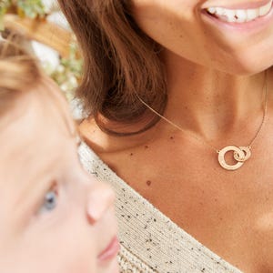 Könnte beinhalten: Eine goldfarbene Halskette mit zwei ineinandergreifenden Kreisen. Die Halskette wird von einer Person getragen und ist vor einem cremefarbenen Pullover zu sehen. Der Hintergrund ist verschwommen, mit Andeutungen von Grün.