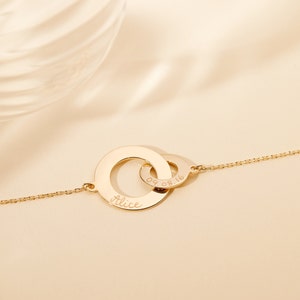 May include: Gold necklace with two interlocking circles. The larger circle is engraved with the name "Alice" and the smaller circle is engraved with the date "09.08.16".