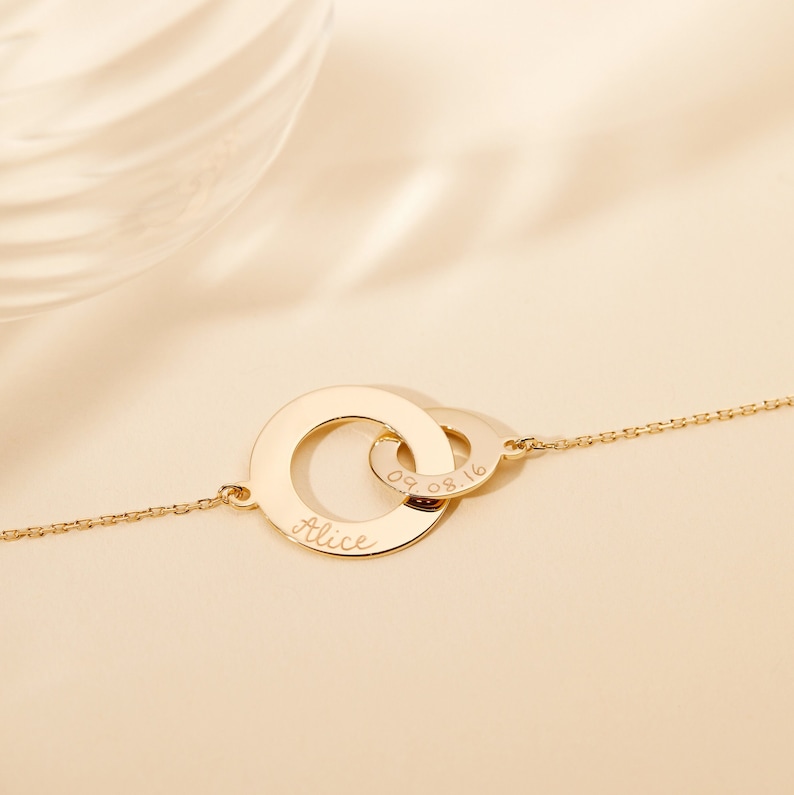 May include: Gold necklace with two interlocking circles. The larger circle is engraved with the name "Alice" and the smaller circle is engraved with the date "09.08.16".