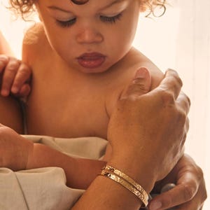 Puede incluir: Dos brazaletes dorados con textura martillada se llevan en un brazo. El brazo lo sostiene otra persona, con un niño en el fondo. Los brazaletes tienen un diseño sencillo y elegante, que refleja la luz.