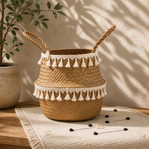 May include: Woven basket with braided handles and white tassel trim. The basket is made of natural fibers and has a round shape. The basket is sitting on a cream-colored rug.