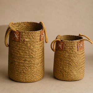 Puede incluir: Dos cestas de paja tejida de diferentes tamaños. Las cestas tienen forma cilíndrica con asas de cuerda y parches de cuero con un diseño en forma de estrella. Las cestas son de color beige natural.