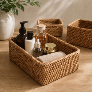 May include: A rectangular woven rattan basket filled with skincare products, including a brown glass bottle with a pump, a white bottle, and a small glass bottle with a dropper. A rolled beige towel and a gold-lidded jar are also inside.