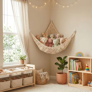 Puede incluir: Una acogedora habitación infantil con una hamaca de macramé llena de peluches. Una estantería de madera exhibe libros y juguetes coloridos. Un mueble de almacenamiento de madera con contenedores se encuentra cerca de una ventana con cortinas transparentes.