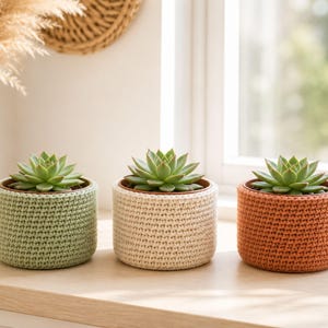 May include: Three small succulent plants in cylindrical pots, each covered with a textured, hand-crocheted cozy. The covers are in solid shades of sage green, cream, and terracotta orange, featuring a consistent, dense stitch pattern.