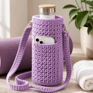 Op de afbeelding: Een lavendelgehaakte waterfleshouder met een riem en een zak met een smartphone. De houder bevat een roestvrijstalen waterfles met een houten deksel. Een opgerolde yogamat en een witte handdoek staan op de achtergrond.
