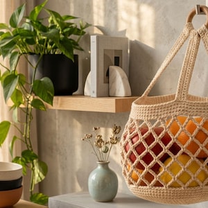 Puede incluir: Una bolsa de red tejida en color beige con un asa de anillo de madera, llena de varias frutas redondas que incluyen naranjas, limones y manzanas rojas, colgada de un gancho de pared junto a un estante con una planta, un libro y un jarrón.