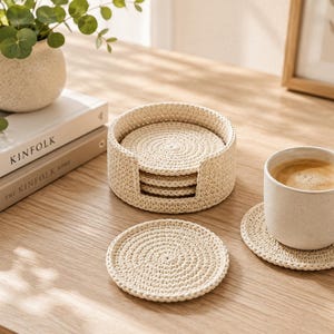 May include: A set of round, cream-colored crocheted coasters with a matching circular holder. The coasters feature a dense, spiral-stitched texture. One coaster is placed flat, another sits under a ceramic mug, and several are stacked inside the holder.
