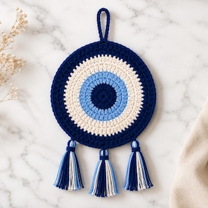 May include: A circular crochet wall hanging featuring a concentric eye pattern in dark blue, light blue, and white yarn. The piece includes a top hanging loop and three decorative tassels at the bottom, each composed of matching dark blue, light blue, and white strands.