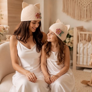 May include: Two matching cream-colored crochet beanies featuring pointed cat ears and a decorative flower appliqué with a pink center and green leaves on the side. The hats are crafted from a fine knit yarn and have a structured, rounded fit.