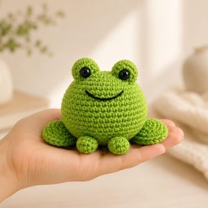 May include: A small, round, bright green crocheted frog amigurumi toy. The frog features a textured yarn surface, two prominent eyes with black pupils, and a simple black stitched smile across its face. It has two small front feet.