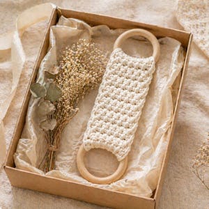 May include: A cream-colored, macrame bath scrub with wooden handles, presented in a brown gift box with dried flowers and tissue paper. The box is tied with a cream-colored ribbon, creating a natural and rustic aesthetic.