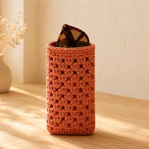 May include: A cylindrical crochet case in a terracotta orange color featuring a repeating shell stitch pattern. The open top of the case holds a pair of dark brown tortoiseshell sunglasses, revealing the textured yarn construction of the holder.