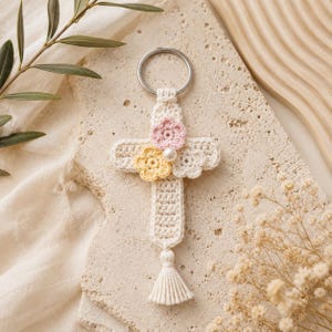 May include: A cream-coloured crochet cross keyring featuring a silver metal ring at the top. The cross is decorated with three small crocheted flowers in pastel pink, yellow, and white, with a single white pearl bead at the centre. A small cream-coloured tassel hangs from the bottom of the cross.