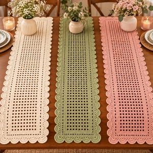 Puede incluir: Tres caminos de mesa rectangulares de ganchillo en color crema, verde salvia y rosa empolvado. Cada camino presenta un patrón de rejilla central con un borde sólido y un acabado festoneado. Están dispuestos uno al lado del otro sobre una mesa.