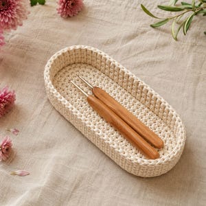 Può includere: Un cestino ovale all'uncinetto color crema contiene due uncinetti di legno. Il cestino è su un tessuto dai toni neutri, con fiori rosa e verde sullo sfondo. Il cestino all'uncinetto è un articolo fatto a mano.