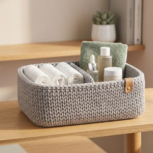 May include: A rectangular gray knitted storage basket with a tan leather tab and gold rivet. The basket features an internal divider, holding three rolled white cloths, a green folded towel, a glass dropper bottle, and two small cosmetic jars.