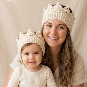 May include: A mother and child are wearing matching cream-coloured crochet crown headbands. The crowns have a textured design with rounded points. The background is a neutral beige fabric. The child is wearing a cream-coloured long-sleeved shirt.