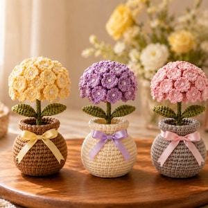 Puede incluir: Tres flores de hortensia de ganchillo hechas a mano en macetas pequeñas. Las flores son amarillas, moradas y rosas, con tallos y hojas verdes. Cada maceta es de un color diferente, con una cinta a juego atada alrededor. Las flores se exhiben en una bandeja de madera.