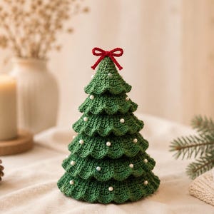 Könnte beinhalten: Ein grüner, gehäkelter Weihnachtsbaum als Dekoration mit gestuften, wellenförmigen Schichten, die mit kleinen weißen perlenartigen Kügelchen verziert sind. Die Spitze des Baumes ist mit einer kleinen, leuchtend roten Häkelschleife versehen.