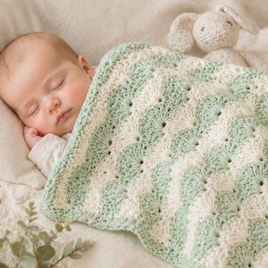 Könnte beinhalten: Ein schlafendes Baby liegt unter einer handgefertigten, gehäkelten Decke. Die Decke hat ein sich wiederholendes Muster aus abwechselnden weißen und hellgrünen Streifen. Ein Stofftier-Hase liegt daneben und verleiht der Szene einen Hauch von Verspieltheit.