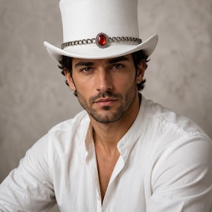 May include: A white top hat featuring a silver-toned metal chain band that encircles the crown. The center of the band is adorned with an oval-shaped, faceted red gemstone set in a decorative silver-toned metal frame with a scalloped edge.