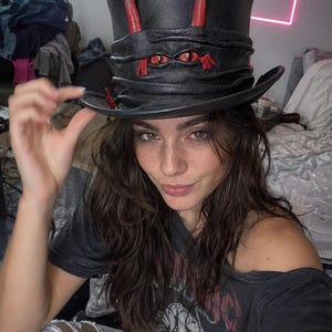 Puede incluir: Un sombrero de copa negro con un acabado texturizado similar al cuero, adornado con dos cuernos erguidos de puntas rojas y un par de ojos rojos realistas incrustados en la banda plegada. El sombrero tiene un ala ancha y una copa estructurada.