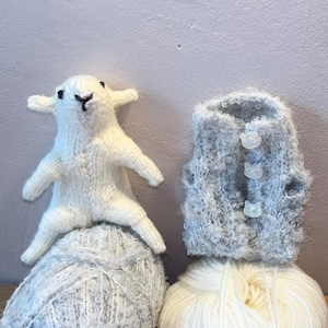 May include: A white knitted lamb figurine sitting on a ball of grey speckled yarn next to a grey fuzzy vest with three small cat-shaped buttons, which is resting on a ball of solid white yarn. The items are arranged side by side.