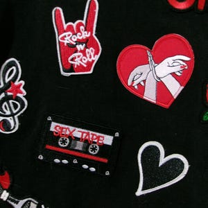 May include: A black fabric surface decorated with various embroidered patches including a red hand gesture with the text Rock n Roll, a red heart with bound hands, a cassette tape labeled SEX TAPE, a treble clef, a red rose, and a black heart.